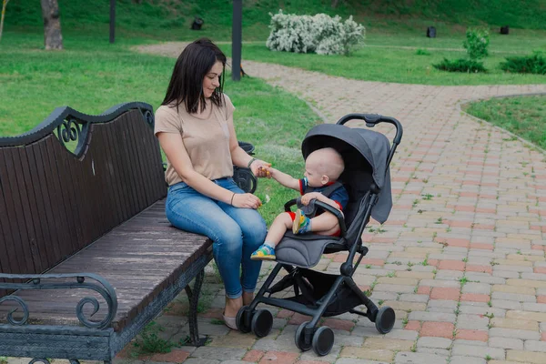 Mooie moeder zit op een bankje met een baby in een kinderwagen op een zonnige dag in de zomer. — Stockfoto