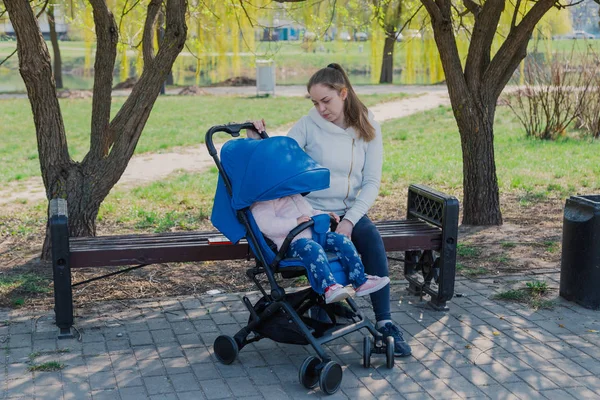 Молодая мать с ребенком в коляске сидит на скамейке в солнечный день . — стоковое фото
