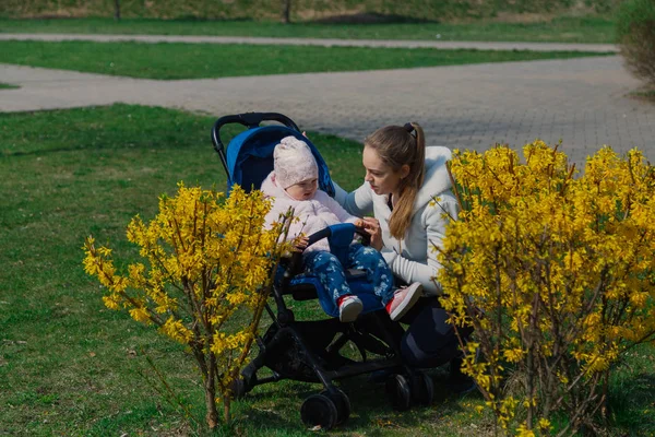 Молодая красивая стильная мама с ребенком в коляске рядом с желтым кустом летом . — стоковое фото