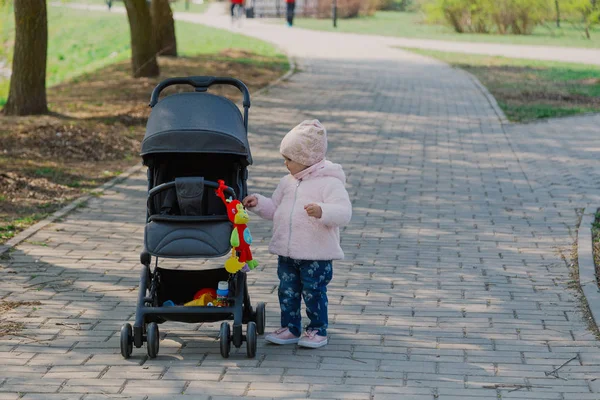 Маленькая девочка катает детскую коляску в парке. Ребенок в парке играет с коляской . — стоковое фото