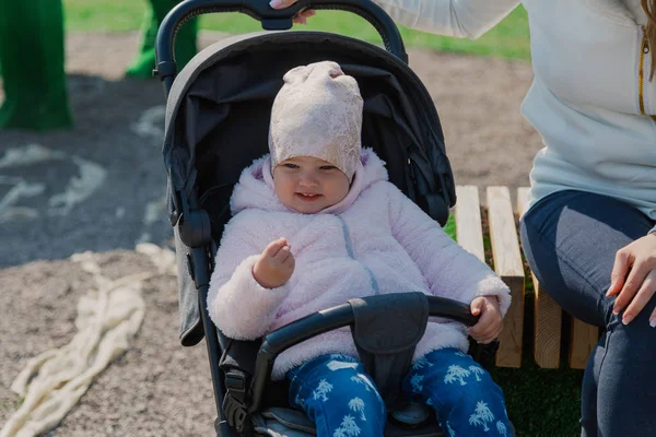 A young mother with a baby in a pram sits on a bench on a sunny day. — Stock Photo, Image