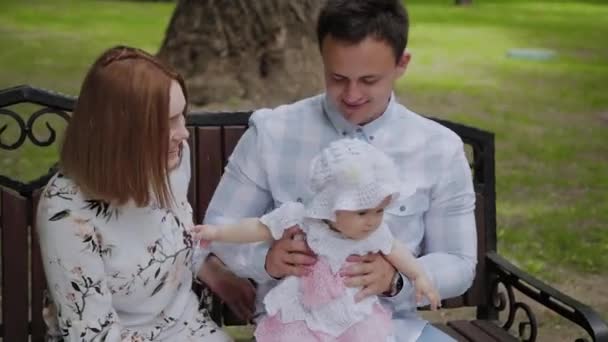 Mladá rodina s jeho synem, spočívající v parku v létě na lavičce. Krásná žena se svým manželem a dítětem sedět na lavičce v pozadí městské krajiny — Stock video