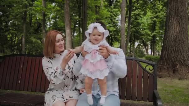 Feliz familia joven con su hijo descansando en el parque en el verano en un banco. Una hermosa mujer con su marido y un niño sentado en un banco en el fondo del paisaje urbano — Vídeo de stock