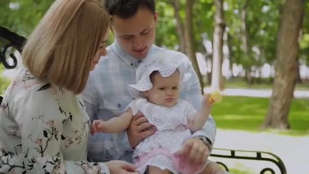 Glad ung familj med hans son vilar i parken i sommar på en bänk. En vacker kvinna med sin man och ett barn som sitter på en bänk i bakgrunden av det urbana landskapet — Stockvideo