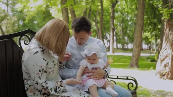 Feliz familia joven con su hijo descansando en el parque en el verano en un banco. Una hermosa mujer con su marido y un niño sentado en un banco en el fondo del paisaje urbano — Vídeos de Stock