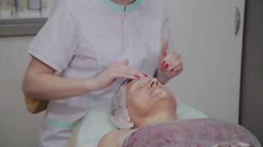 Kadın kozmetolojist kozmetoloji kliniğinde yaşlı kadına yüz masajı yapıyor.