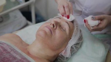 Güzellik uzmanı, bir kozmetoloji kliniğinde yaşlı bir kadının yüzünü yıkama işlemini gerçekleştirir.