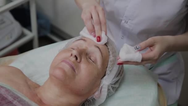 Esteticista realiza o procedimento de lavar o rosto de uma mulher idosa em uma clínica de cosmetologia . — Vídeo de Stock