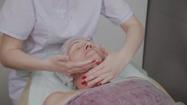 Cosmetóloga femenina haciendo masaje facial a anciana en clínica cosmetológica . — Vídeos de Stock