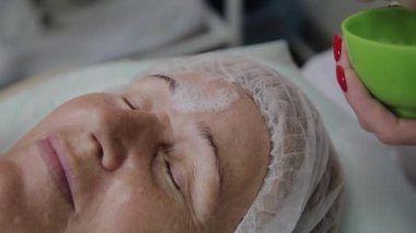 Kadın kozmetolojist, kozmetoloji kliniğinde yaşlı kadına yüz maskesi uyguluyor.