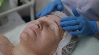 Kadın kozmetolojist kozmetoloji kliniğinde yaşlı kadına yüz masajı yapıyor.