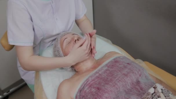 여성 미용사, 미용클리닉에서 노인 여성에게 얼굴 마사지. — 비디오