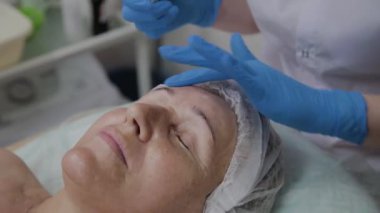 Profesyonel kozmetolojist güzellik salonunda yaşlı bir kadının yüzüne serum uygular.