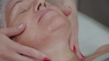 Kadın kozmetolojist kozmetoloji kliniğinde yaşlı kadına yüz masajı yapıyor.