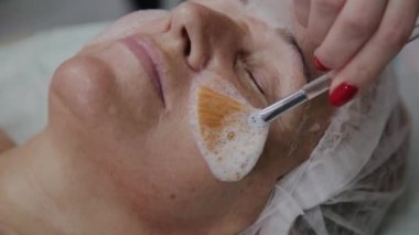 Kadın kozmetolojist, kozmetoloji kliniğinde yaşlı kadına yüz maskesi uyguluyor.