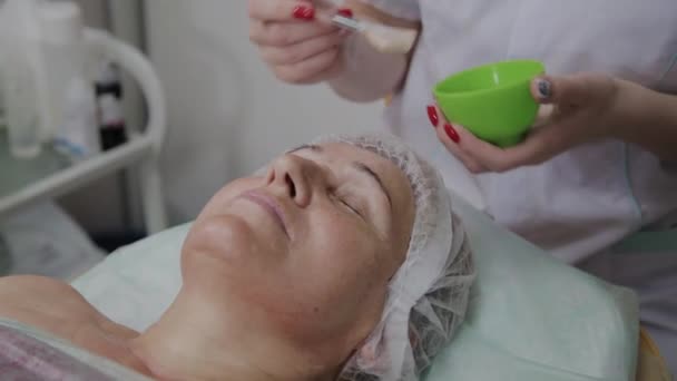 Cosmetologist fêmea aplica máscara facial à mulher idosa na clínica cosmetológica . — Vídeo de Stock