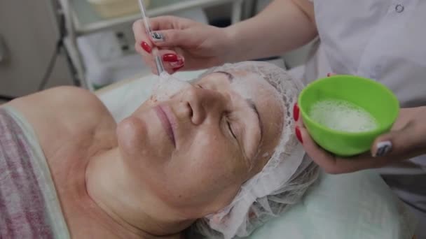 Cosmetologist fêmea aplica máscara facial à mulher idosa na clínica cosmetológica . — Vídeo de Stock