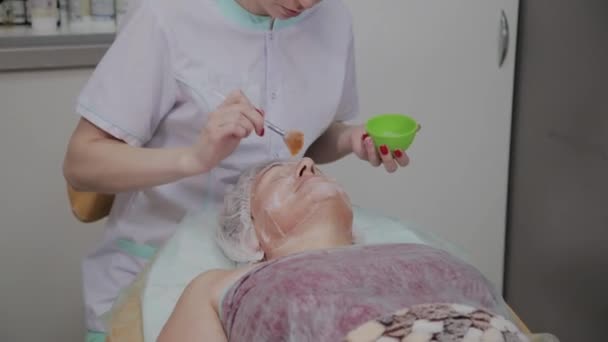 Γυναικεία καλλυντικός χρησιμοποιεί μάσκα προσώπου για την ηλικιωμένη γυναίκα σε καλλυντικολογική κλινική. — Αρχείο Βίντεο