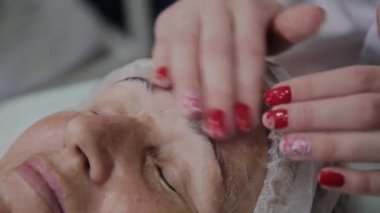 Kadın kozmetolojist kozmetoloji kliniğinde yaşlı kadına yüz masajı yapıyor.
