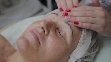 Kadın kozmetolojist kozmetoloji kliniğinde yaşlı kadına yüz masajı yapıyor.