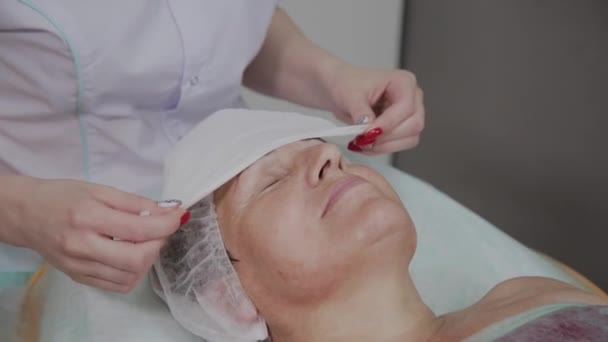 Esteticista profissional remove toalhetes do rosto de uma mulher idosa no escritório de cosmetologia . — Vídeo de Stock