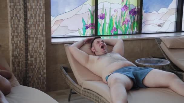 Hombre con sobrepeso tumbado en una tumbona junto a la piscina cubierta . — Vídeo de stock