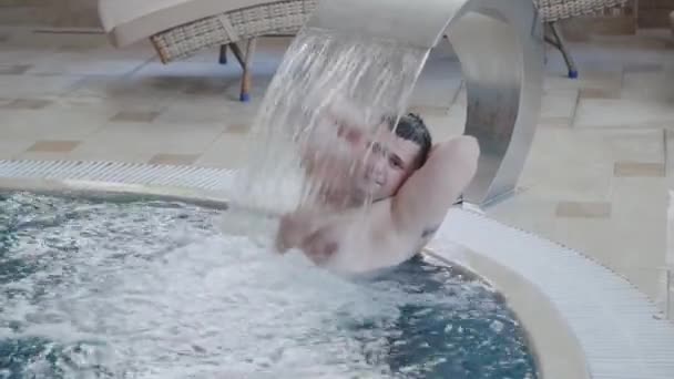 Fröhlicher Mann schwimmt unter einem Wasserfall und posiert im Pool. — Stockvideo