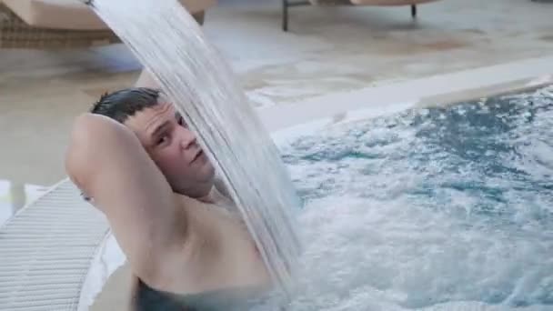 Vrolijke man zwemt onder een waterval en poses in het zwembad. — Stockvideo