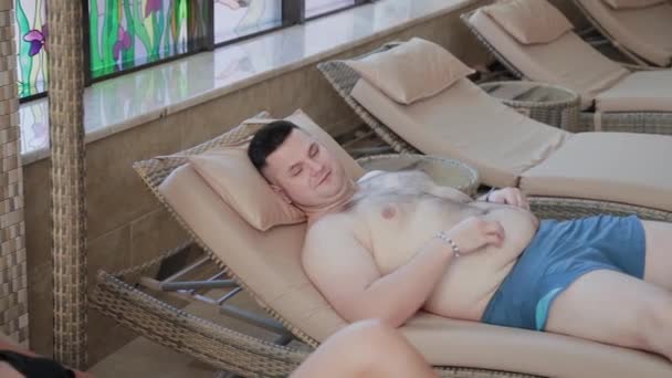 太り過ぎの男と美しい女性は、覆われたスイミングプールのラウンジャーに横たわっています. — ストック動画
