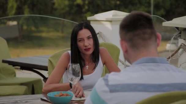 En vacker kvinna och en man sitter vid ett bord i en restaurang på en öppen terrass. — Stockvideo