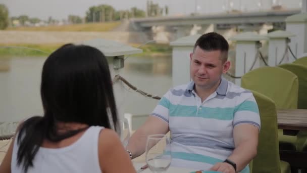 Uma mulher bonita e um homem estão sentados em uma mesa em um restaurante em um terraço aberto . — Vídeo de Stock