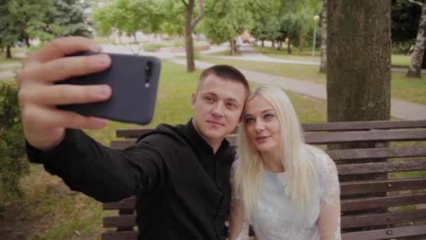 Pareja feliz enamorada sentada en un banco en el parque de la ciudad y haciendo selfie por teléfono . — Vídeo de stock