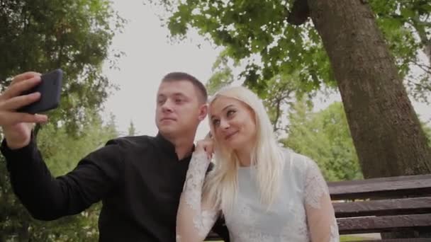 Ευτυχισμένο ζευγάρι στην αγάπη κάθονται σε ένα παγκάκι στο πάρκο της πόλης και να κάνει selfie στο τηλέφωνο. — Αρχείο Βίντεο