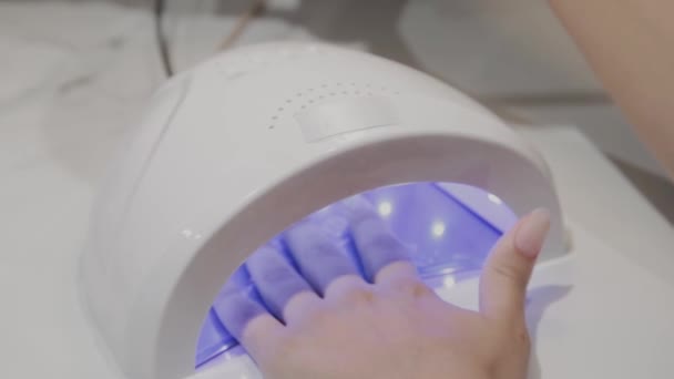 Una chica joven seca esmalte de uñas bajo una lámpara ultravioleta en el salón de manicura . — Vídeos de Stock