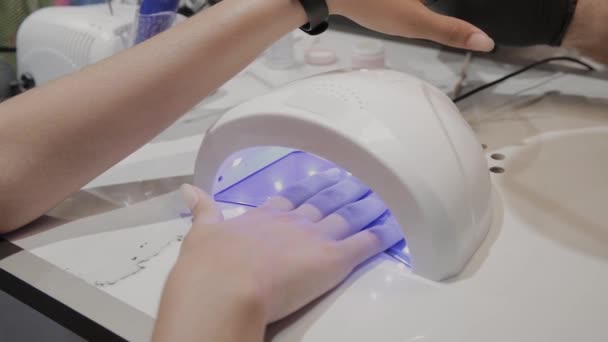 Uma jovem seca esmalte sob uma lâmpada ultravioleta no salão de manicure . — Vídeo de Stock