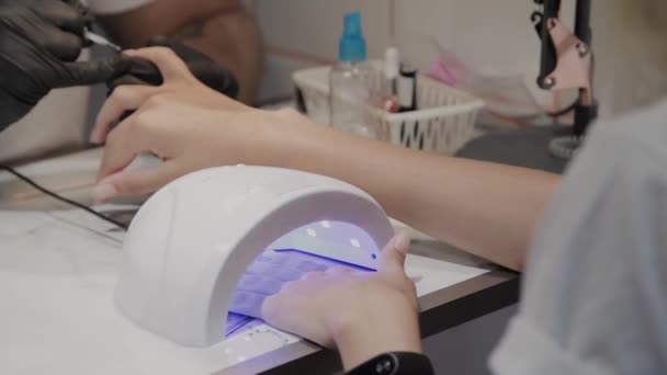 Młoda dziewczyna wysycha lakier do paznokci pod lampą ultrafioletowego w salonie manicure. — Wideo stockowe