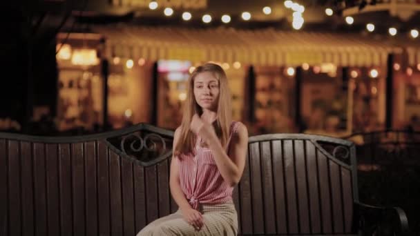 Menina bonita em um banco à noite em um parque . — Vídeo de Stock