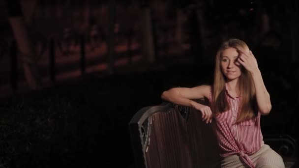 Mooi jong meisje op een bankje in de avond in een park. — Stockvideo