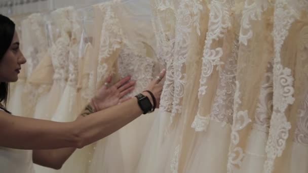 Hermosa chica elige un vestido de novia en un salón de bodas, un hombre se esconde en vestidos . — Vídeos de Stock