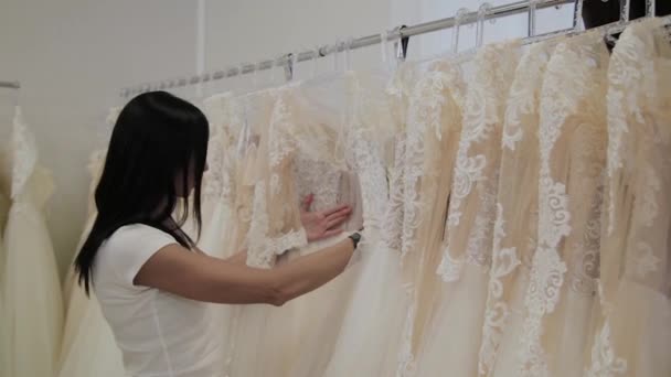 Piękna dziewczyna wybiera suknię ślubną w salonie ślubnej, człowiek ukrywa się w sukienkach. — Wideo stockowe