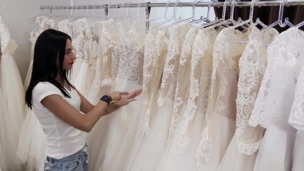 Piękna dziewczyna wybiera suknię ślubną w salonie ślubnej, człowiek ukrywa się w sukienkach. — Wideo stockowe