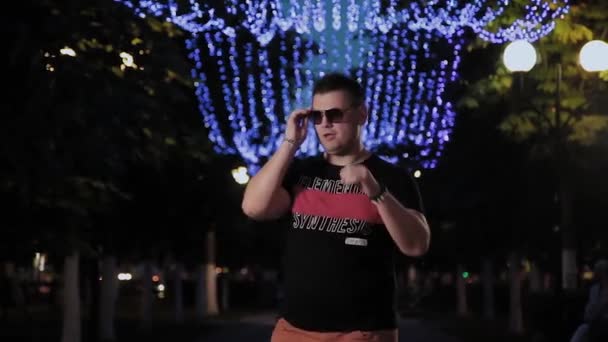 Pozytywny i zabawny człowiek w okulary przeciwsłoneczne wieczorem na alei miasta. — Wideo stockowe