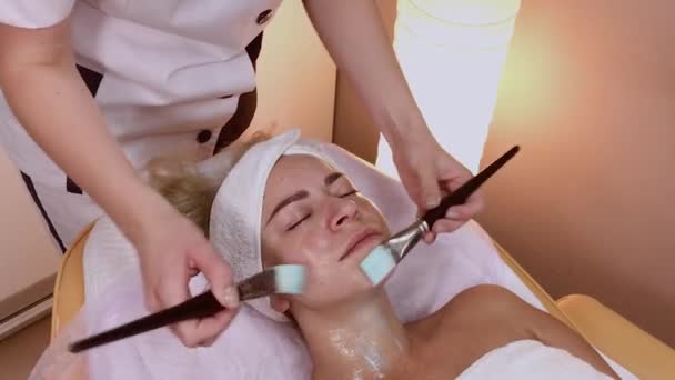 Профессиональный косметолог наносит маску с кисточками на лицо женщины . — стоковое видео