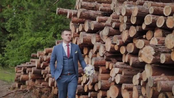 Knappe bruidegom in een jasje en met een boeket staat in de buurt van een stapel logs. — Stockvideo