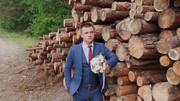 Beau marié dans une veste et avec un bouquet se tient près d'une pile de bûches . — Video