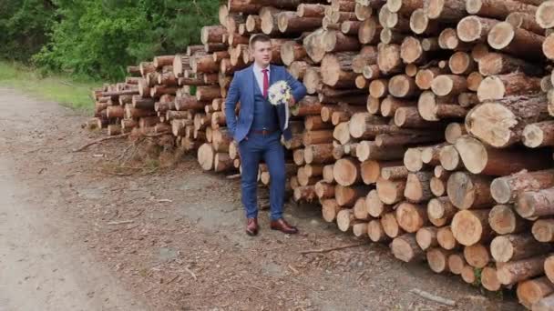 Bonito noivo em uma jaqueta e com um buquê fica perto de uma pilha de troncos . — Vídeo de Stock