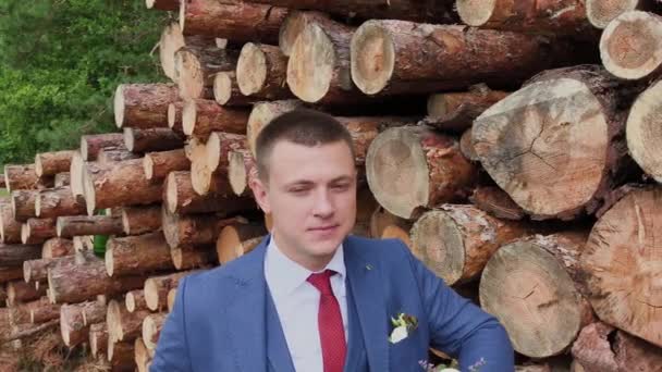 Bonito noivo em uma jaqueta e com um buquê fica perto de uma pilha de troncos . — Vídeo de Stock