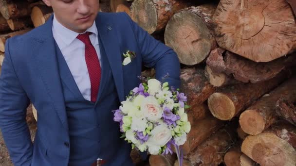 Beau marié dans une veste et avec un bouquet se tient près d'une pile de bûches . — Video