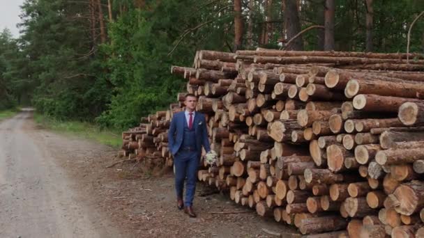 Novio guapo en una chaqueta y con un ramo de pie cerca de una pila de troncos . — Vídeos de Stock