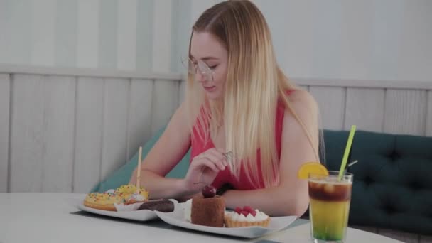 Vacker ung flicka sitter vid ett bord i ett kafé med en tallrik desserter. — Stockvideo